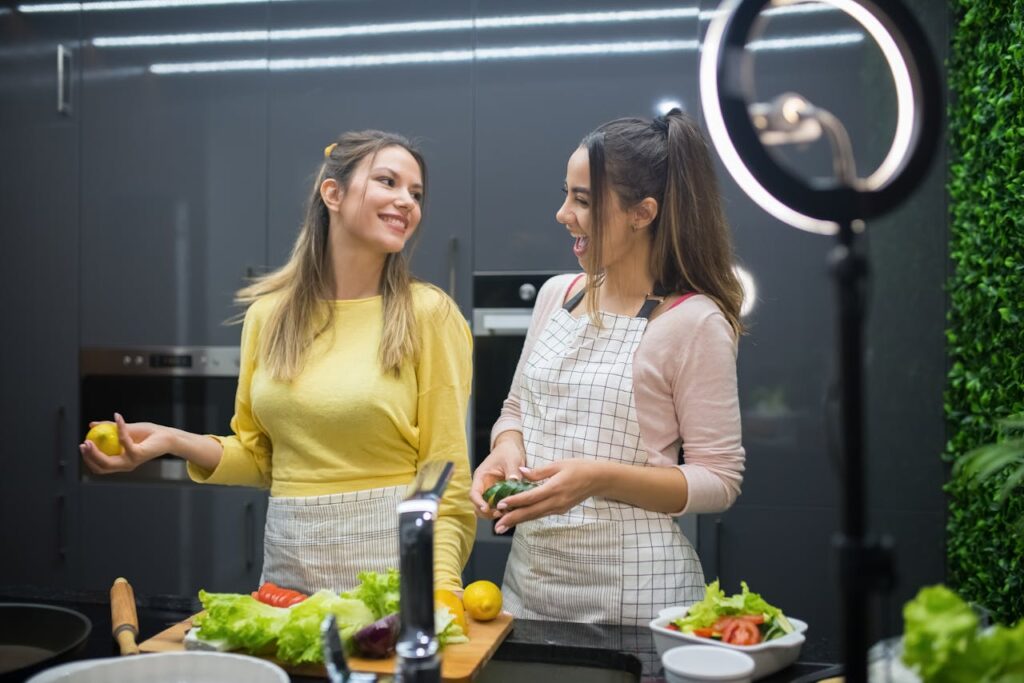 Two women enjoying cooking with fresh vegetables under bright kitchen lighting.